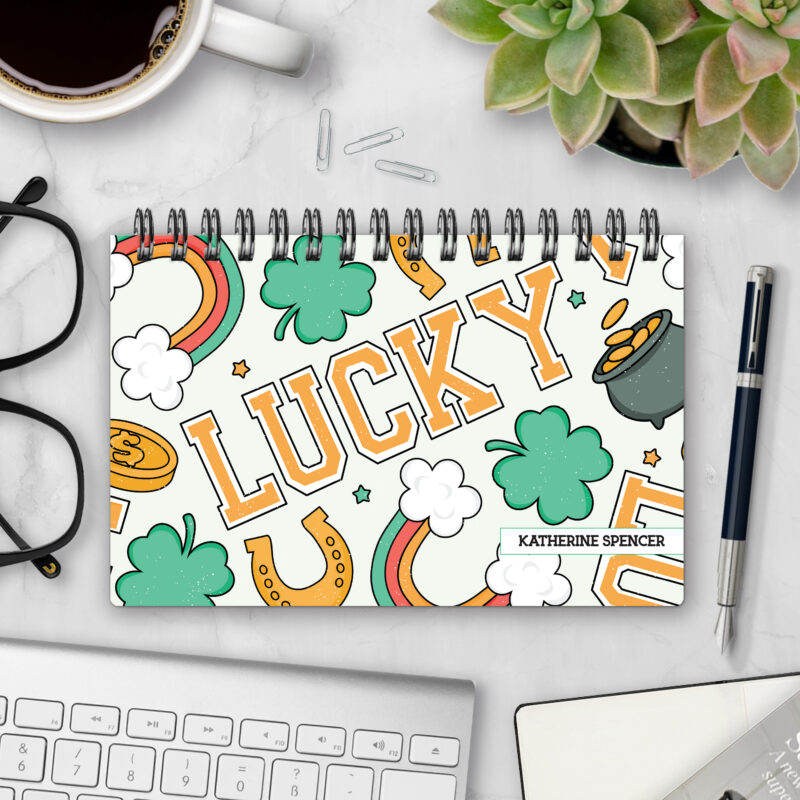 Top-bound landscape planner with a Lucky cover featuring shamrocks, horseshoes, rainbows, and a pot of gold around bold "LUCKY" varsity lettering on a cream background, styled on a marble desk with coffee, succulents, glasses, and a pen.