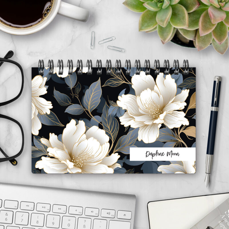 Top-bound landscape planner with a Midnight Bloom cover featuring oversized ivory peonies with gold linework and slate-blue foliage on a black background, styled on a marble desk with coffee, succulents, glasses, and a pen.