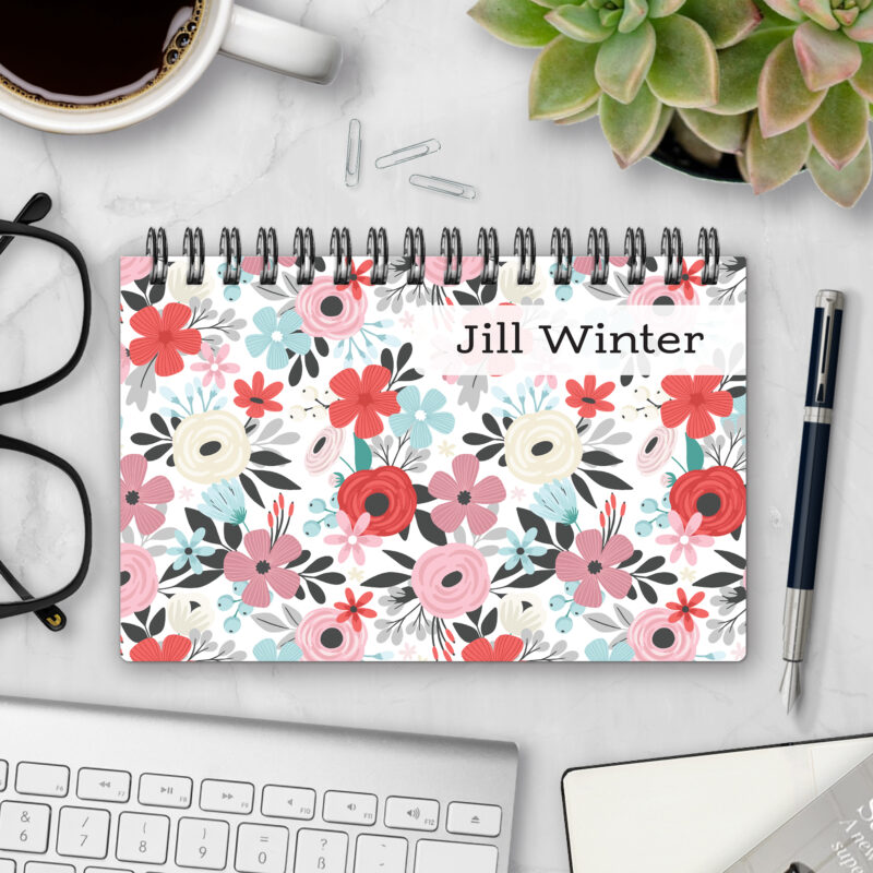 Top-bound landscape planner with a Petal Pusher cover featuring pink, coral, and cream roses with charcoal foliage and pale blue accents on a white background, styled on a marble desk with coffee, succulents, glasses, and a pen.