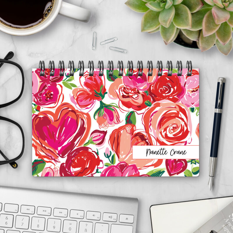 Top-bound landscape planner with a Painted Love cover featuring loose, expressive brushstroke roses and heart-shaped blooms in shades of red, pink, coral, and peach with vibrant green leaves on a white background, styled on a marble desk with coffee, succulents, glasses, and a pen.