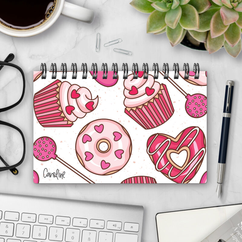 Top-bound landscape planner with a Sugar Rush cover featuring pink cupcakes, heart-sprinkled donuts, cake pops, and chocolate-covered hearts on a cream background, styled on a marble desk with coffee, succulents, glasses, and a pen.