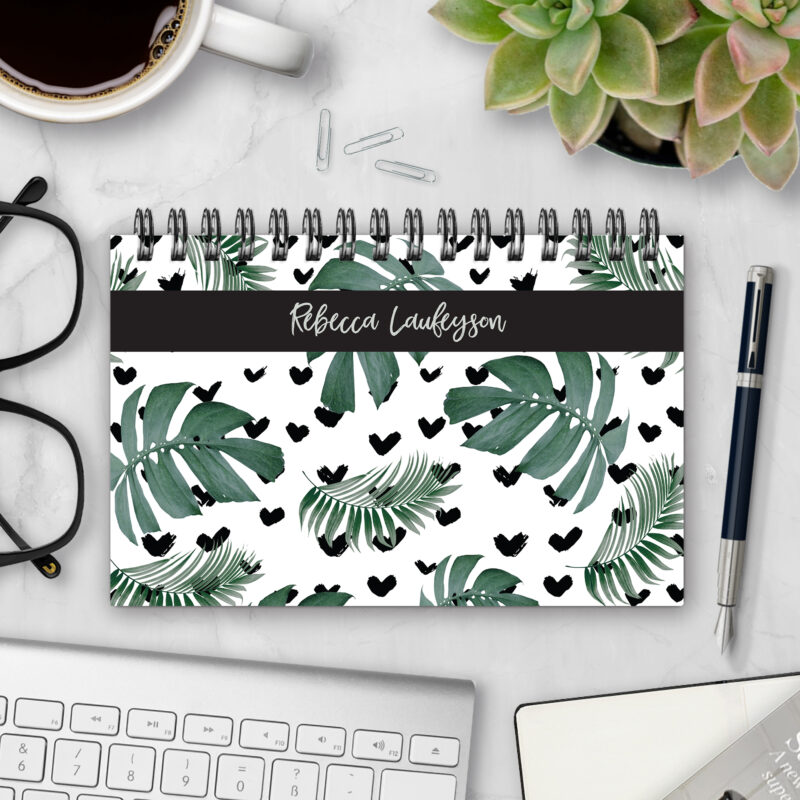 Top-bound landscape planner with a Tropical Love cover featuring monstera and palm leaves with scattered black brushstroke hearts on a white background, styled on a marble desk with coffee, succulents, glasses, and a pen.