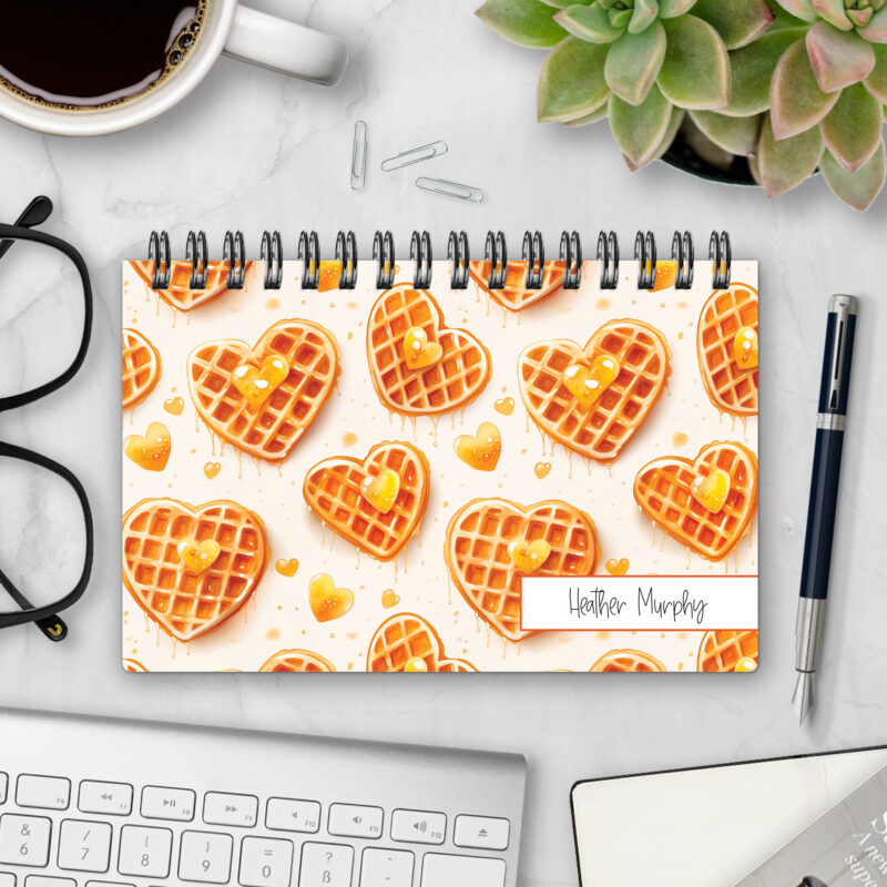 Top-bound landscape planner with a Slow Mornings cover featuring heart-shaped waffles drizzled with golden syrup and topped with heart-shaped pats of butter on a cream background, styled on a marble desk with coffee, succulents, glasses, and a pen.