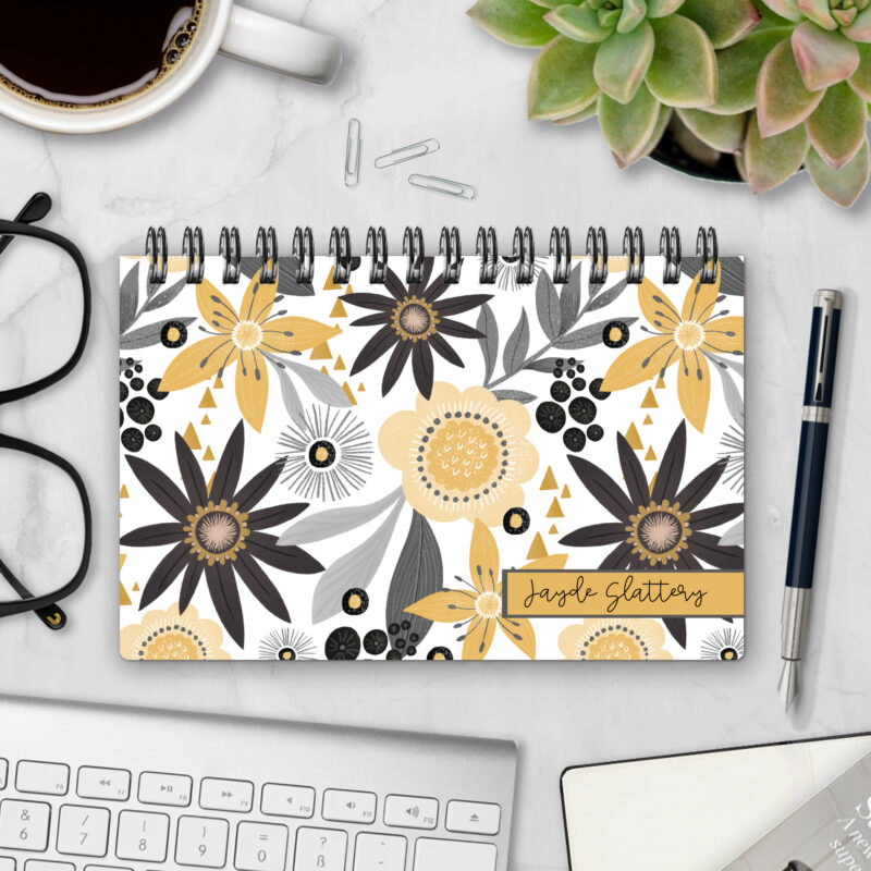 Top-bound landscape planner with a Sunglow cover featuring large mustard-yellow, charcoal, and gray stylized flowers with delicate foliage on a white background, styled on a marble desk with coffee, succulents, glasses, and a pen.