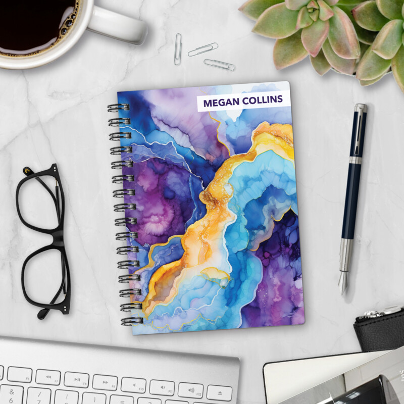 Personalized hardcover notebook in Amethyst Geode design with purple, blue, and gold marbled cover styled on marble desk with coffee, glasses, and pen