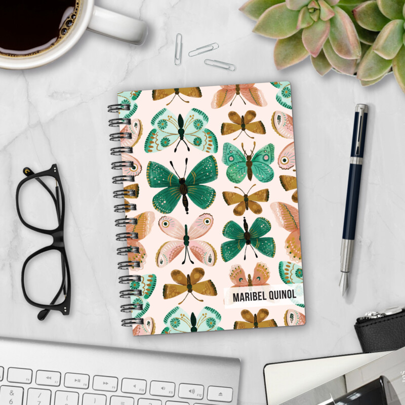 Personalized hardcover notebook in Flutter By design with painted butterflies in green, pink, and gold on cream background styled on marble desk with pen