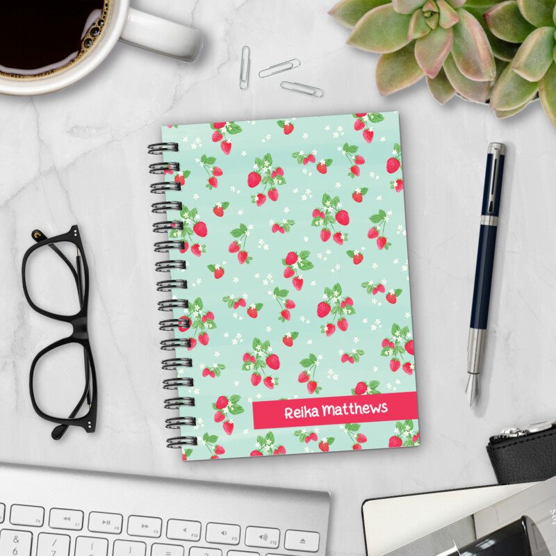 Personalized hardcover notebook in Berry Overbooked design with red strawberries and white blossoms on mint green background styled on marble desk
