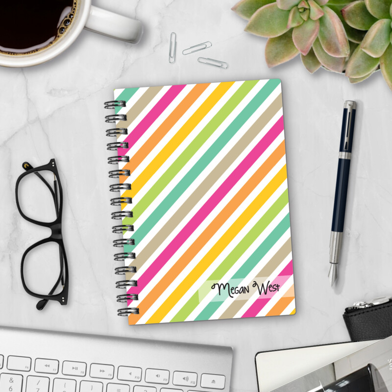 Personalized hardcover notebook in Stripe Hype design with diagonal rainbow stripes in pink, orange, yellow, green, and teal styled on marble desk with pen