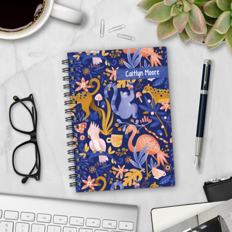Personalized hardcover notebook in Jungle Love design with whimsical monkeys, leopards, flamingos, and cockatoos on navy background styled with pen