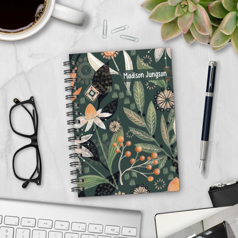 Personalized hardcover notebook in Night Garden design with botanical leaves, berries, and folk florals on dark green background styled on marble desk