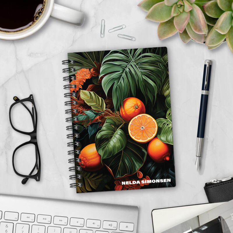 Personalized hardcover notebook in Blood Orange Jungle design with oranges and tropical monstera leaves on dark background styled on marble desk with pen