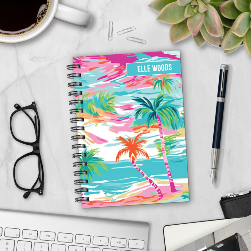 Personalized hardcover notebook in Aquarelle Sky design with watercolor palm trees in pink, turquoise, and orange styled on marble desk with coffee and pen