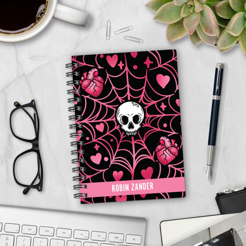 Personalized hardcover notebook in Love Bites design with white skull, anatomical hearts, and pink spider webs on black background styled with pen