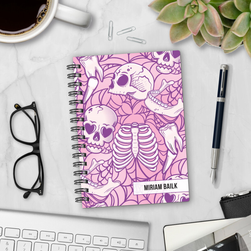 Personalized hardcover notebook in Lovesick Bones design with pink skulls, ribcage, bones, and heart eyes on purple spider web background styled with pen