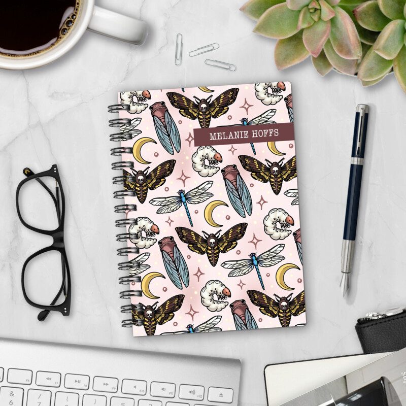Personalized hardcover notebook in Moonlit Macabre design with death moths, cicadas, dragonflies, grubs, and crescent moons on pink background styled with pen