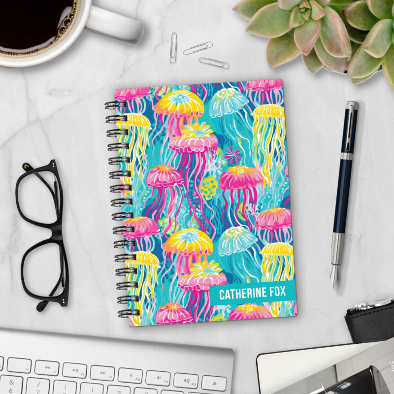 Personalized hardcover notebook in Neon Jellyfish design with bright pink, yellow, and turquoise jellyfish on blue ocean background styled with pen