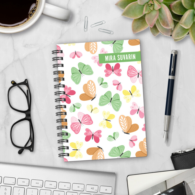 Personalized hardcover notebook in Whimsical Wings design with cheerful pink, green, yellow, and orange butterflies on white background styled with pen