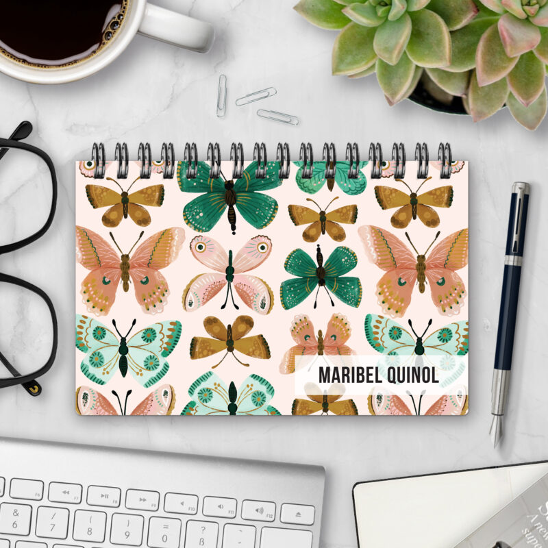 Top-bound landscape planner with a Flutter By cover featuring rows of folk-art butterflies in coral, mustard, mint, and emerald on a soft blush background, styled on a marble desk with coffee, succulents, glasses, and a pen.