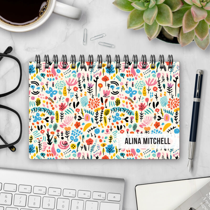 Top-bound landscape planner with a Garden Graffiti cover featuring playful hand-drawn flowers, leaves, and abstract marks in pink, turquoise, yellow, and black on a cream background, styled on a marble desk with coffee, succulents, glasses, and a pen.