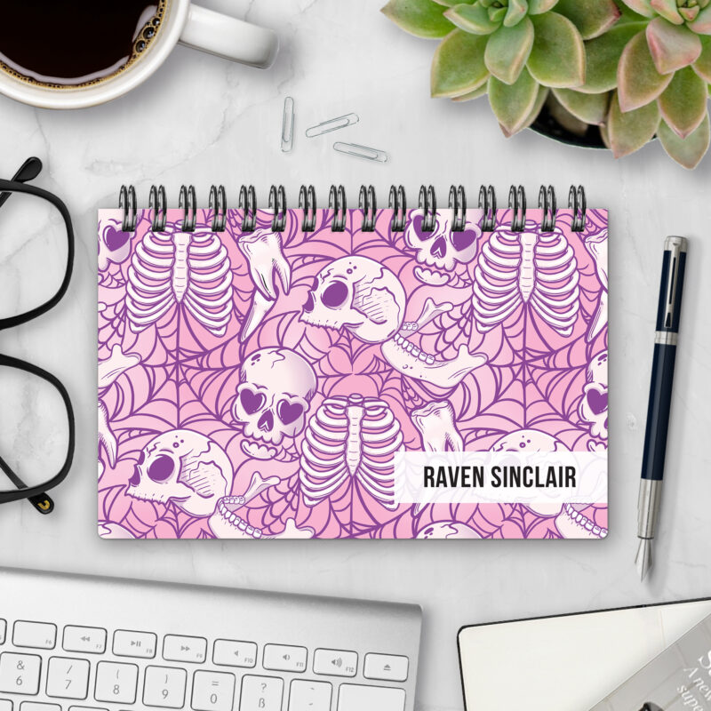 Top-bound landscape planner with a Lovesick Bones cover featuring pastel pink and lavender skulls with heart-shaped eyes, ribcages, and spiderwebs, styled on a marble desk with coffee, succulents, glasses, and a pen.
