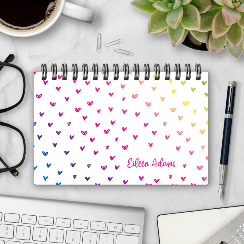 Top-bound landscape planner with a Rainbow Hearts cover featuring scattered hand-drawn hearts in a rainbow gradient from blue and purple to pink, orange, and yellow on a white background, styled on a marble desk with coffee, succulents, glasses, and a pen.