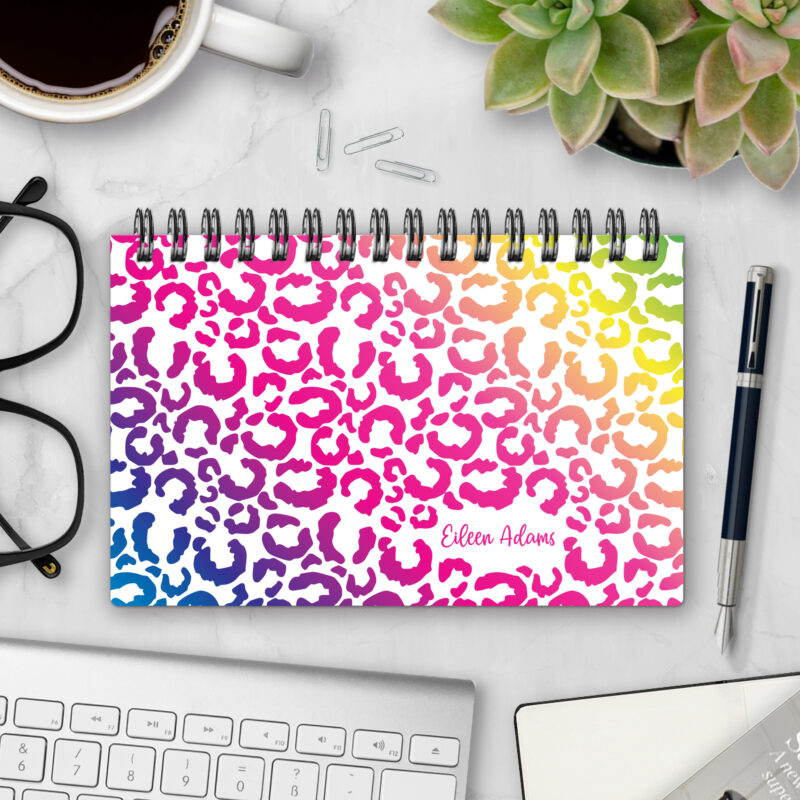 Top-bound landscape planner with a Rainbow Leopard cover featuring leopard-print spots in a vibrant rainbow gradient from blue and purple through pink, orange, and yellow on a white background, styled on a marble desk with coffee, succulents, glasses, and a pen.