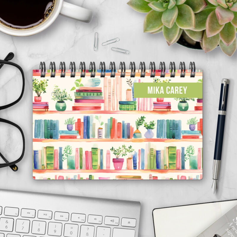 Top-bound landscape planner with a Shelf Control cover featuring watercolor illustrations of bookshelves filled with colorful books and potted plants, styled on a marble desk with coffee, succulents, glasses, and a pen.