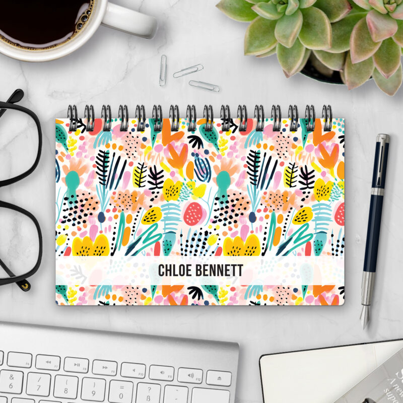 Top-bound landscape planner with a Summer Splash cover featuring an abstract painted pattern of tropical leaves, dots, and brushstrokes in coral, yellow, teal, pink, and black on a cream background, styled on a marble desk with coffee, succulents, glasses, and a pen.