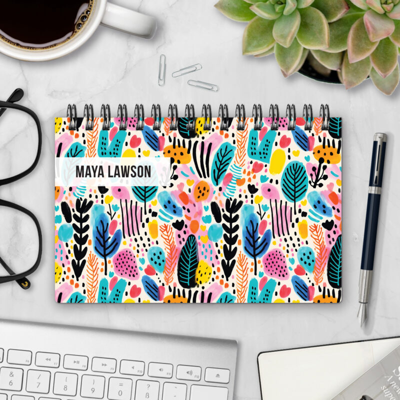 Top-bound landscape planner with a Wild Strokes cover featuring a bold abstract pattern of painted leaves, dots, and brushstrokes in pink, turquoise, yellow, orange, and black on a cream background, styled on a marble desk with coffee, succulents, glasses, and a pen.