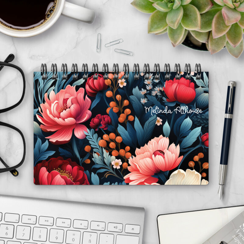 Top-bound landscape planner with a Dark Jewels cover featuring richly colored coral, red, and cream peonies among deep teal foliage on a near-black background, styled on a marble desk with coffee, succulents, glasses, and a pen.