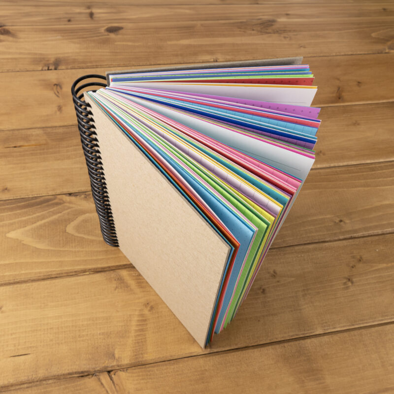 Handmade 5 x 8 inch junk journal with chipboard covers and black plastic coil binding standing open to show colorful mixed paper pages in pink, blue, green, purple, and yellow in assorted printed patterns, displayed on a rustic wood surface. Physical paper journal only, binder and accessories not included. Junk journal is made in the USA by CityGirl Planners