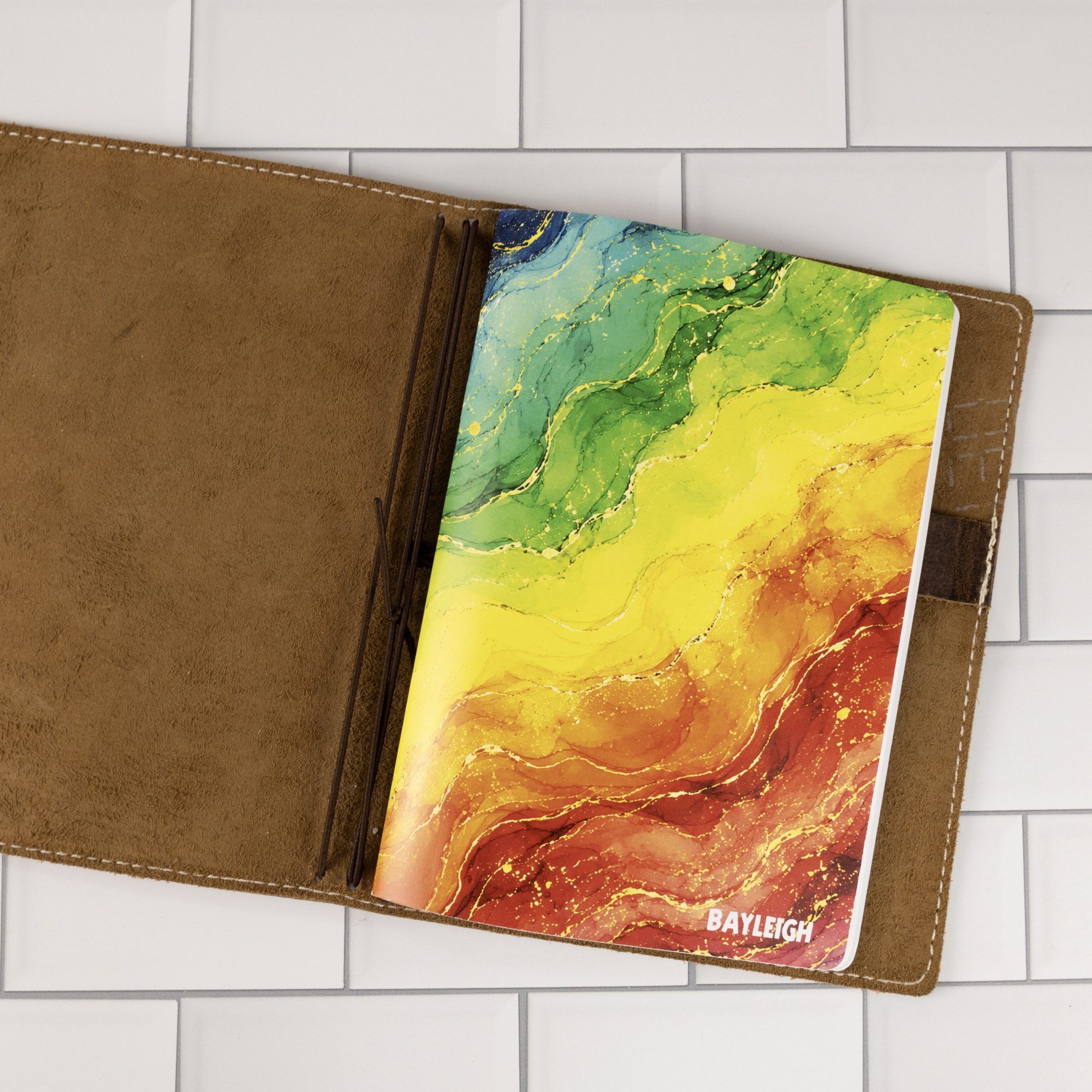 Prism personalized traveler's notebook insert placed atop a closed dark leather traveler's notebook binder, rainbow watercolor marble cover showing.