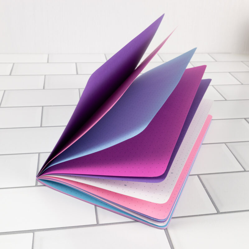 Unicorn Tears traveler's notebook insert fanned open showing layered pages in purple, periwinkle, fuchsia, and pink with dot grid pattern.