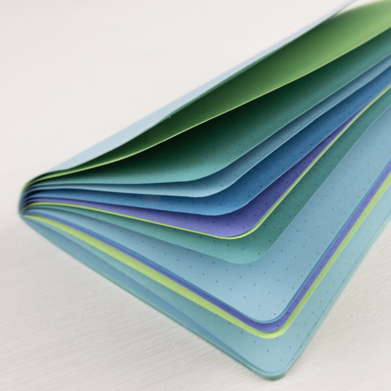 Mermaid Song traveler's notebook insert with colored pages fanned open showing alternating shades of pastel blue, lime green, teal, lavender, and sky blue with dot grid pattern visible on paper edges.
