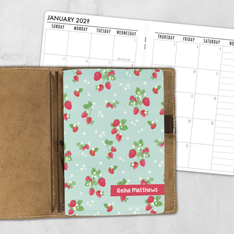 A5 Berry Overbooked lifestyle personalized traveler's notebook refill showing the cover design inside a leather traveler's notebook cover, with a monthly calendar spread visible behind.