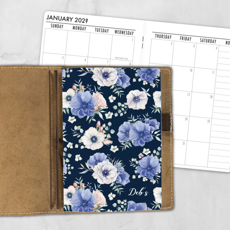 A5 Blooming Mystery lifestyle personalized traveler's notebook refill showing the cover design inside a leather traveler's notebook cover, with a monthly calendar spread visible behind.