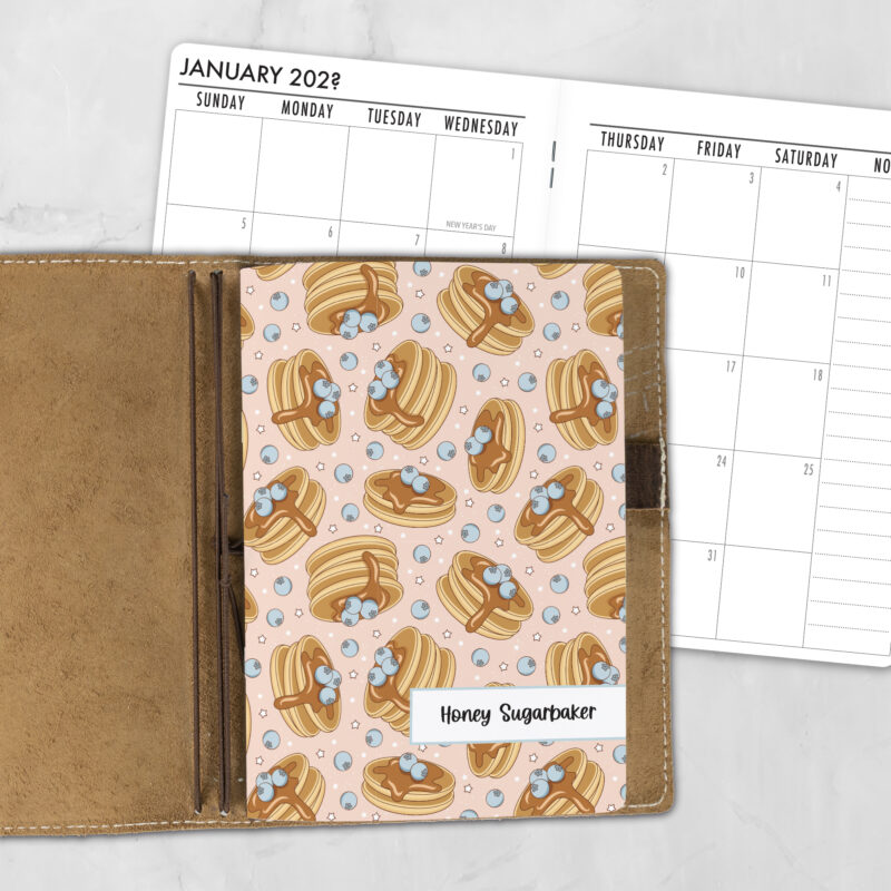 A5 Blueberry Pancakes personalized traveler's notebook refill showing the cover design inside a leather traveler's notebook cover, with a monthly calendar spread visible behind.