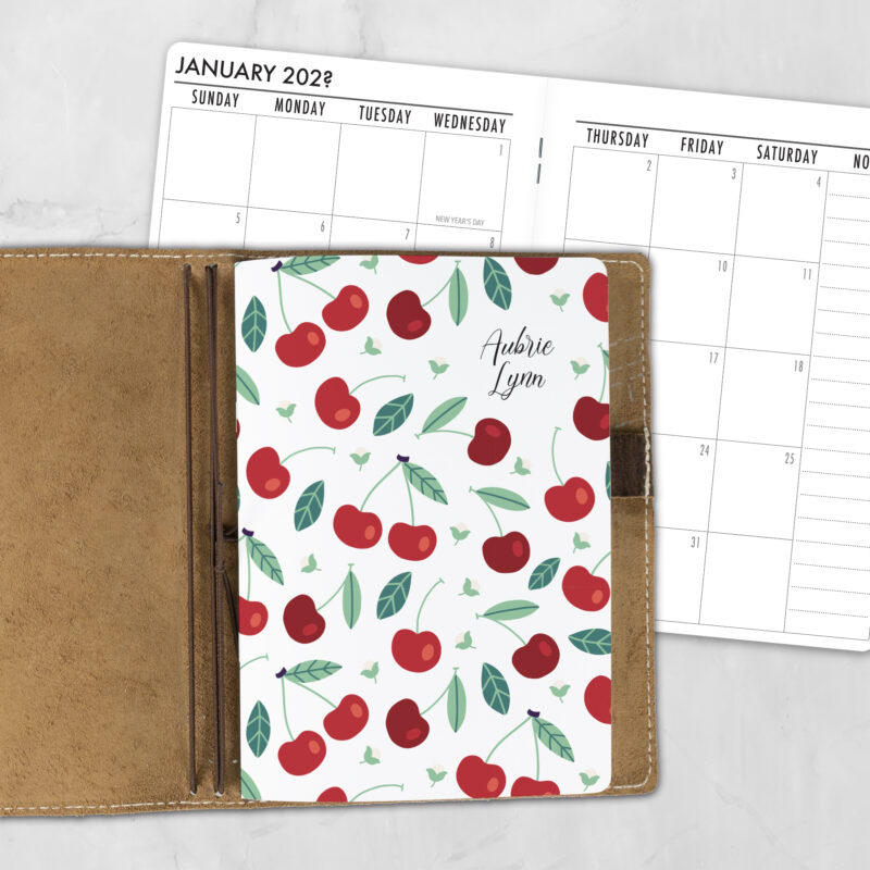 A5 Cherry Sweet personalized traveler's notebook refill showing the cover design inside a leather traveler's notebook cover, with a monthly calendar spread visible behind.