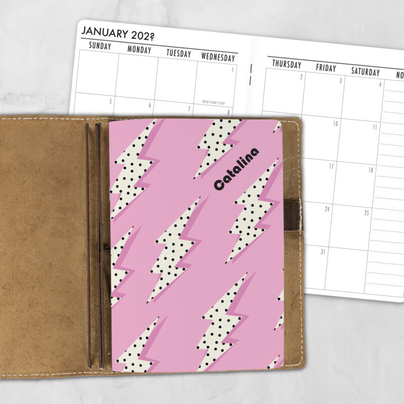 A5 Electric Candy personalized traveler's notebook refill showing the cover design inside a leather traveler's notebook cover, with a monthly calendar spread visible behind.