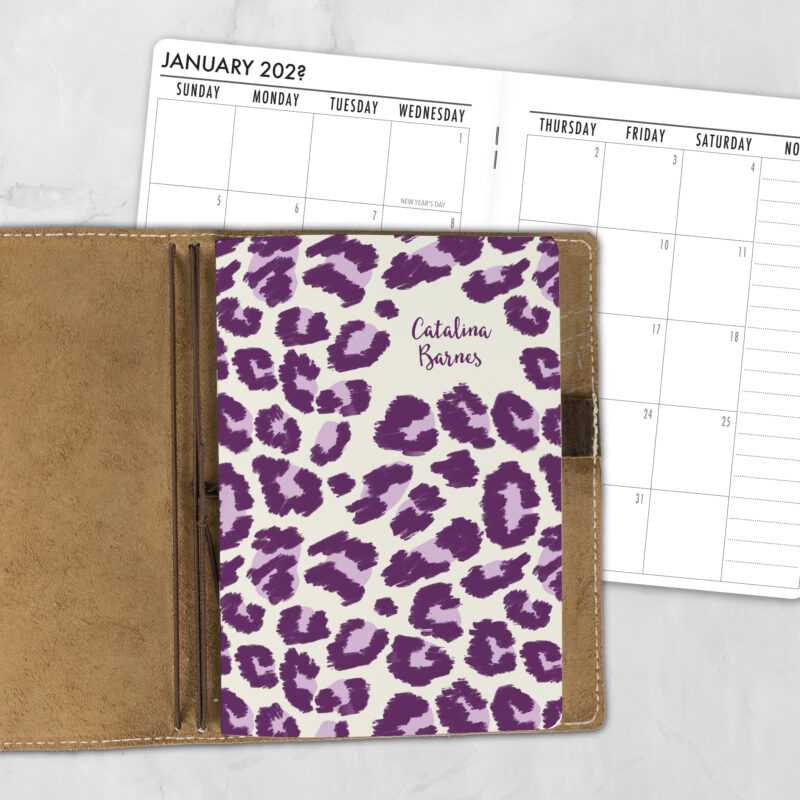A5 Electric Prowl personalized traveler's notebook refill showing the cover design inside a leather traveler's notebook cover, with a monthly calendar spread visible behind.