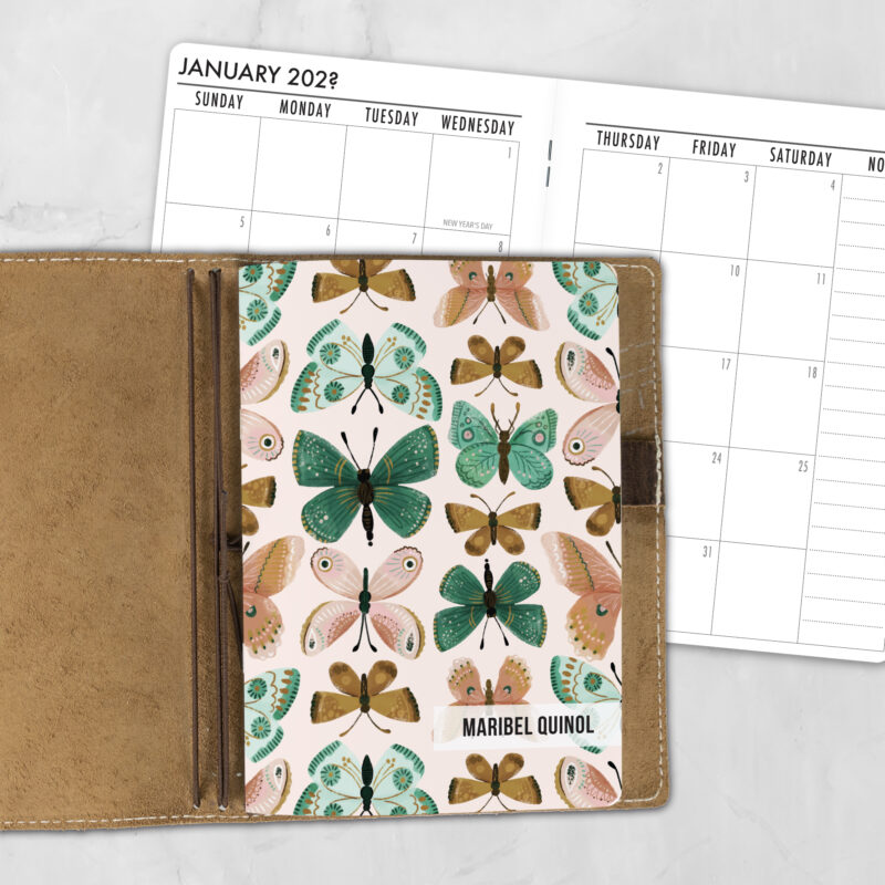 A5 Flutter By lifestyle personalized traveler's notebook refill showing the cover design inside a leather traveler's notebook cover, with a monthly calendar spread visible behind.