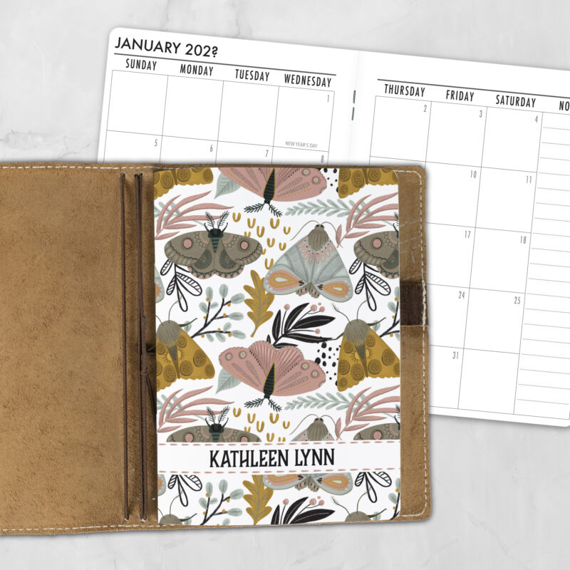 A5 Folkwing personalized traveler's notebook refill showing the cover design inside a leather traveler's notebook cover, with a monthly calendar spread visible behind.