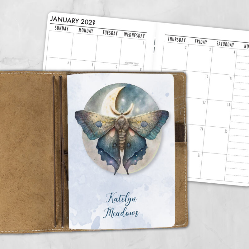 A5 Golden Wings personalized traveler's notebook refill showing the cover design inside a leather traveler's notebook cover, with a monthly calendar spread visible behind.