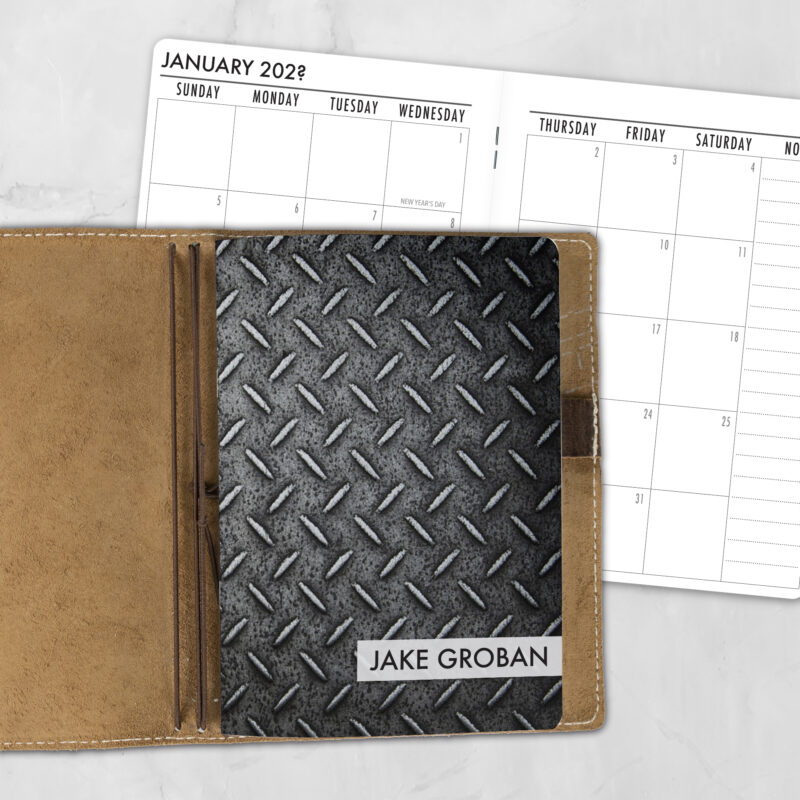 A5 Industrial Strength personalized traveler's notebook refill showing the cover design inside a leather traveler's notebook cover, with a monthly calendar spread visible behind.
