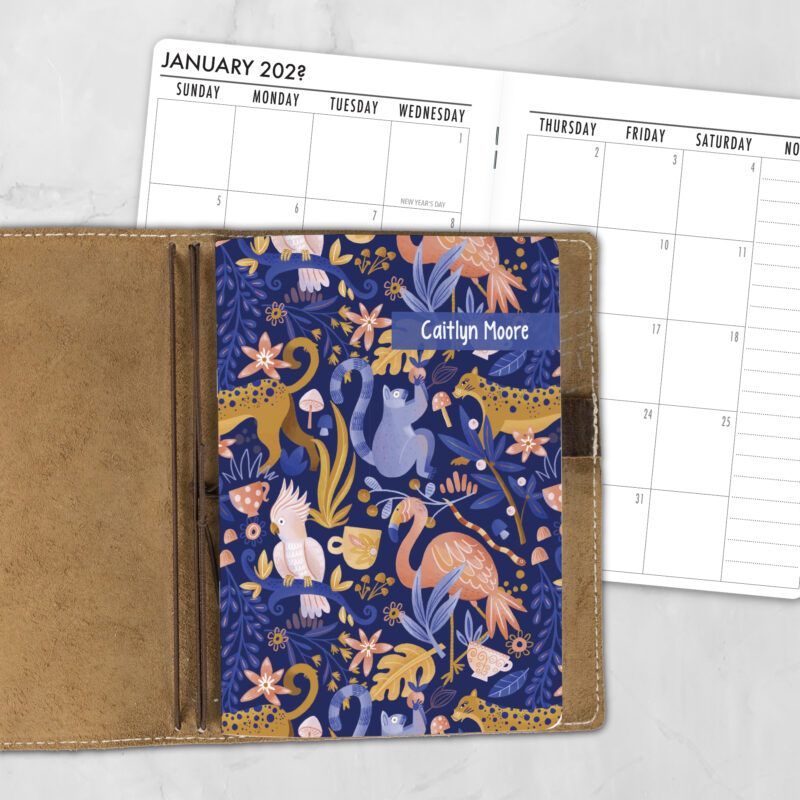 A5 Jungle Love lifestyle personalized traveler's notebook refill showing the cover design inside a leather traveler's notebook cover, with a monthly calendar spread visible behind.
