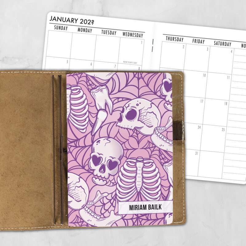 A5 Lovesick Bones lifestyle personalized traveler's notebook refill showing the cover design inside a leather traveler's notebook cover, with a monthly calendar spread visible behind.