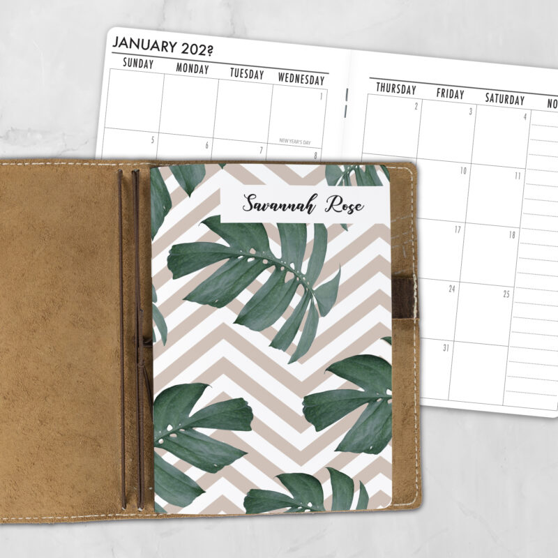 A5 Monstera Summer personalized traveler's notebook refill showing the cover design inside a leather traveler's notebook cover, with a monthly calendar spread visible behind.