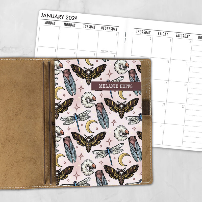 A5 Moonlit Macabre lifestyle personalized traveler's notebook refill showing the cover design inside a leather traveler's notebook cover, with a monthly calendar spread visible behind.