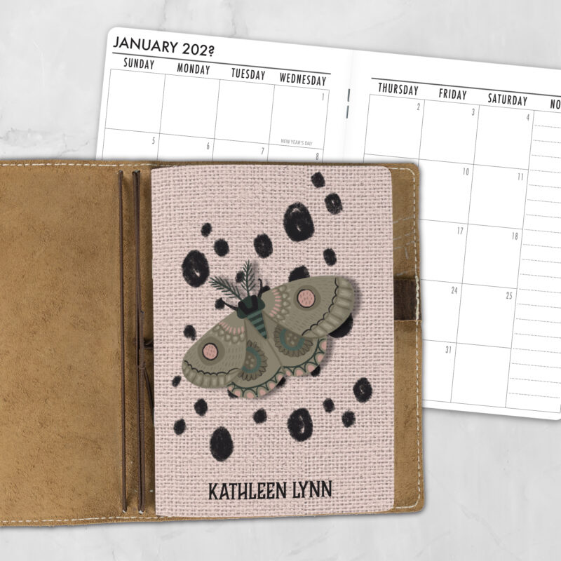 A5 Moth Dust Dot personalized traveler's notebook refill showing the cover design inside a leather traveler's notebook cover, with a monthly calendar spread visible behind.