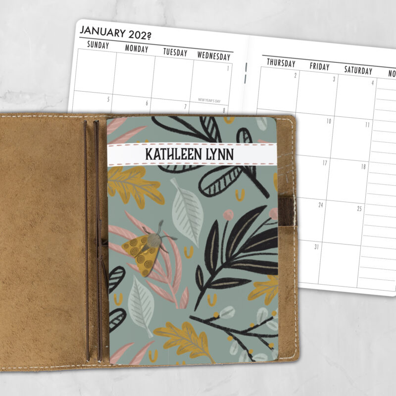 A5 Moth Fern personalized traveler's notebook refill showing the cover design inside a leather traveler's notebook cover, with a monthly calendar spread visible behind.